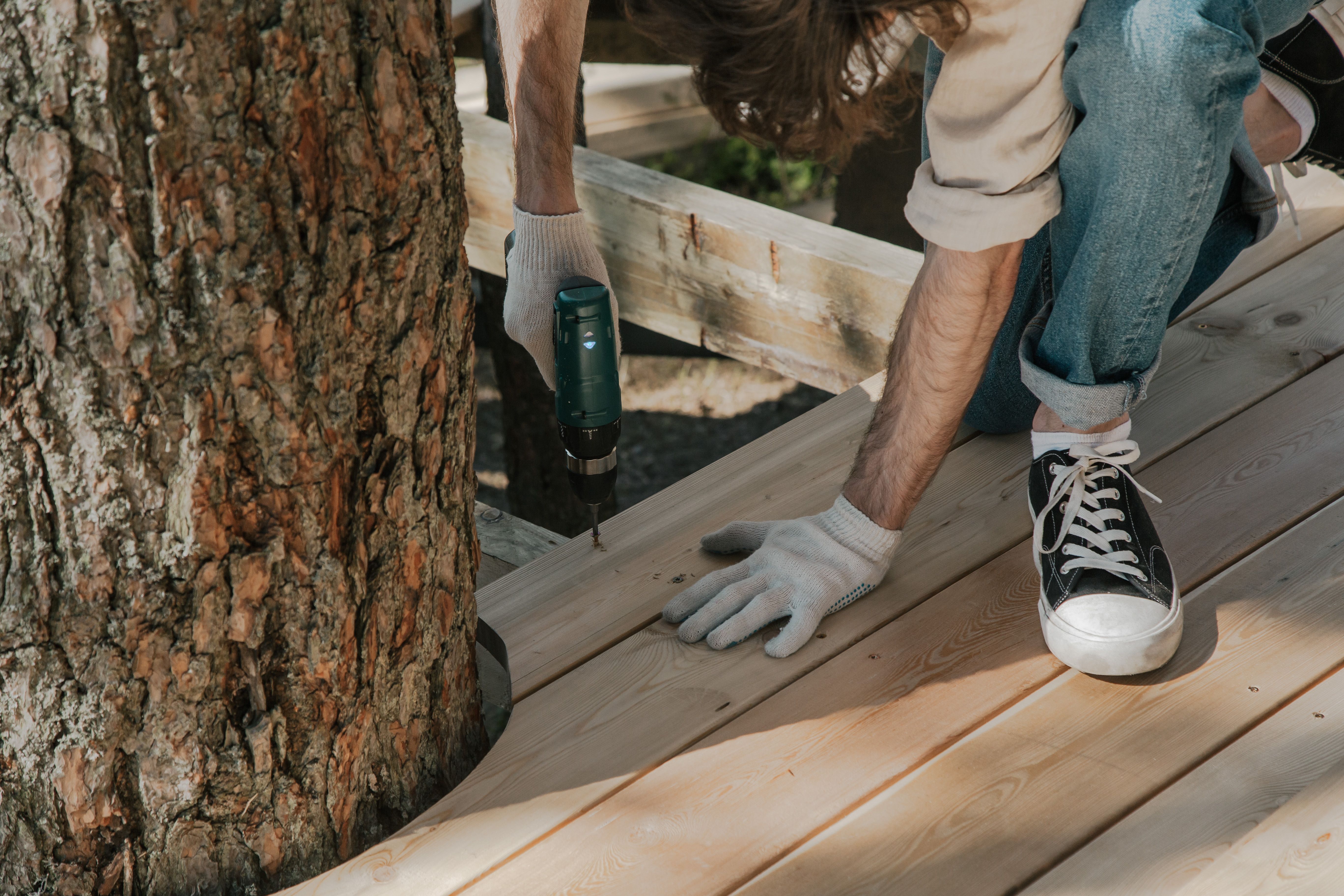 Worker drilling decking boards beside a tree during an outdoor deck construction project